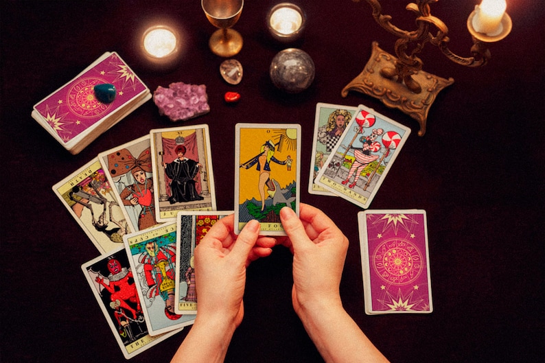 May include: A tarot card reading spread with a variety of cards laid out on a dark purple surface. The cards are surrounded by candles, crystals, and a gold candlestick. The person is holding the "The Fool" card, which depicts a figure standing on a cliff with a staff and a backpack.