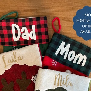 May include: Four personalized Christmas stockings with different patterns and names. The stockings are red, green, and white with black and white plaid patterns. The names on the stockings are "Dad", "Mom", "Laila", and "Mila".