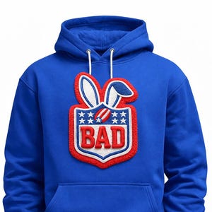 May include: Royal blue hoodie with a large, embroidered patch featuring bunny ears and the word "BAD" in red and white. The patch is framed by a red border and stars, creating a playful design.