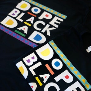May include: Two black t-shirts with the phrase "Dope Black Dad" printed in white letters with colourful accents. The shirts are folded on top of each other.