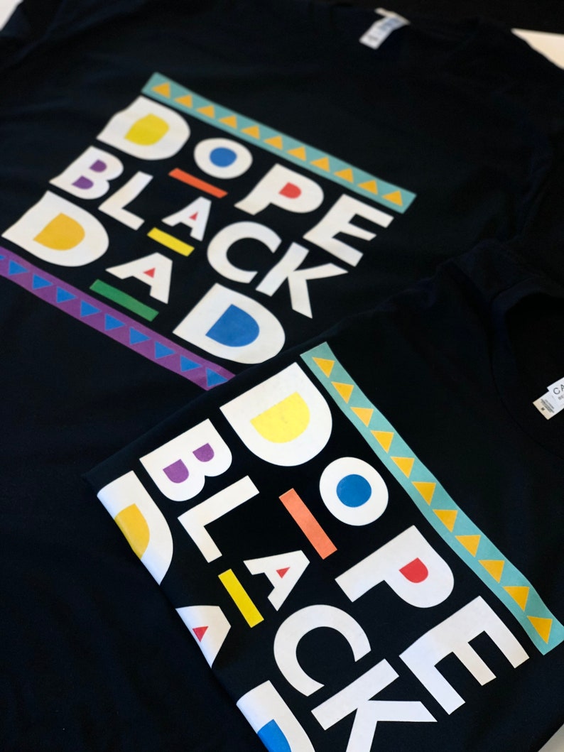 May include: Two black t-shirts with the phrase "Dope Black Dad" printed in white letters with colourful accents. The shirts are folded on top of each other.