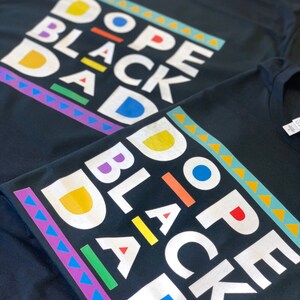 May include: Two black t-shirts with the text "Dope Black Dad" printed in white with colourful accents. The text is in a blocky, retro font.