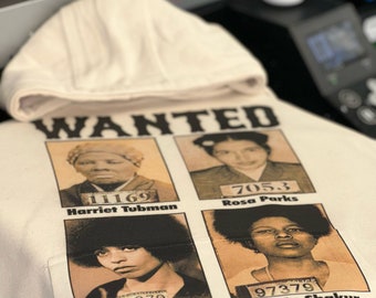 May include: A beige hoodie with a black and white print of four women's mugshots. The mugshots are labeled with the names Harriet Tubman, Rosa Parks, Angela Davis, and Assata Shakur. The text "WANTED" is at the top of the print. The text "WELL-BEHAVED WOMEN SELDOM MAKE HISTORY" is at the bottom of the print.