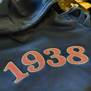 Puede incluir: Una sudadera azul marino con el n&uacute;mero "1938" bordado en rojo y blanco en la parte delantera.