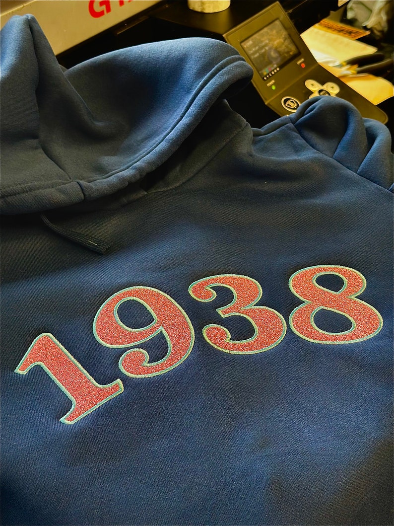 Puede incluir: Una sudadera azul marino con el n&uacute;mero "1938" bordado en rojo y blanco en la parte delantera.
