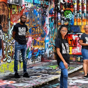May include: Four people stand in front of a graffiti-covered wall, wearing black t-shirts with the words "Black Lives Matter" printed in white. They are all looking at the camera. The wall is covered in colorful spray paint and has a variety of different designs.