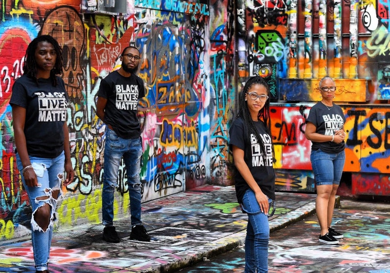 May include: Four people stand in front of a graffiti-covered wall, wearing black t-shirts with the words "Black Lives Matter" printed in white. They are all looking at the camera. The wall is covered in colorful spray paint and has a variety of different designs.