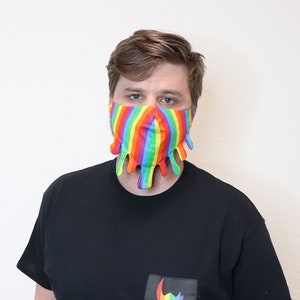 May include: A person wearing a black t-shirt with a rainbow-colored graphic on the pocket. The person is wearing a rainbow-colored face mask with dripping edges.