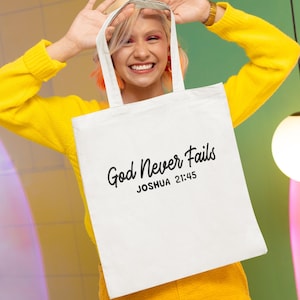 May include: A white tote bag featuring the words "God Never Fails" and "Joshua 21:45" in black script. The bag is held by its handles and has a minimalist design.