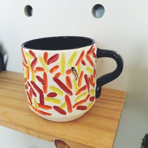Puede incluir: Una taza de cerámica blanca con asa negra. La taza está decorada con un patrón colorido de líneas rojas, amarillas y naranjas.