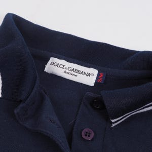 May include: Close-up of a navy blue polo shirt with a white collar and trim. The shirt features a button-down placket with a purple button and a label that reads "DOLCE & GABBANA Intimo". The size tag reads "S/M".