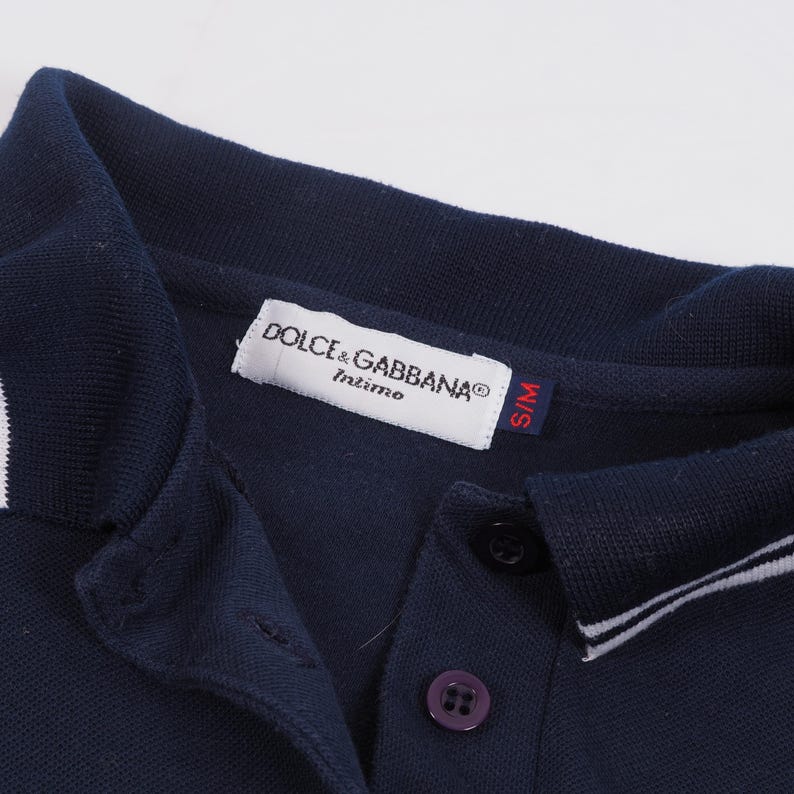 May include: Close-up of a navy blue polo shirt with a white collar and trim. The shirt features a button-down placket with a purple button and a label that reads "DOLCE & GABBANA Intimo". The size tag reads "S/M".