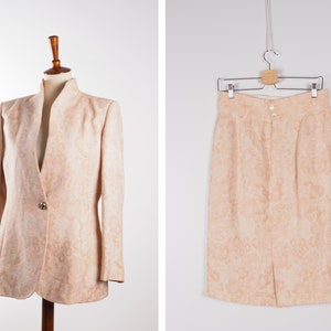 Puede incluir: Un conjunto de blazer y falda de color melocotón con un estampado floral. La chaqueta tiene un cierre de un solo botón y un cuello mandarín. La falda es de largo hasta la rodilla y tiene una silueta recta.