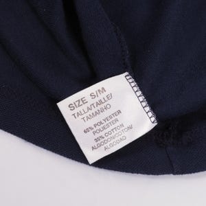 May include: Close-up of a navy blue garment with a white tag. The tag reads "SIZE S/M" and lists the fabric composition as 65% polyester and 35% cotton. The garment's texture is visible.