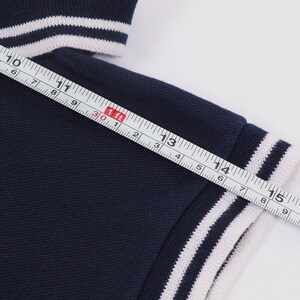 May include: Navy blue polo shirt sleeve with white stripes on the cuff. The sleeve is measured with a ruler, showing a length of approximately 6 inches.