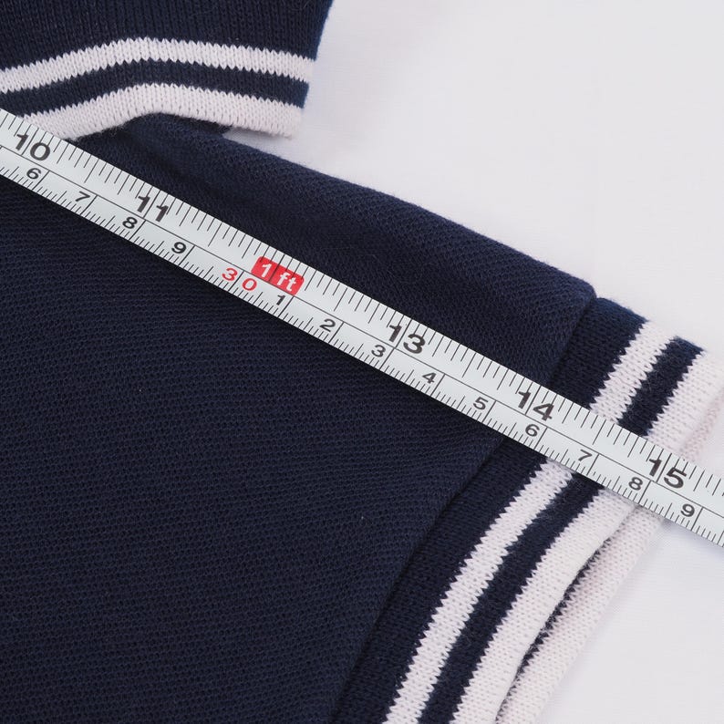 May include: Navy blue polo shirt sleeve with white stripes on the cuff. The sleeve is measured with a ruler, showing a length of approximately 6 inches.