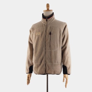 May include: A beige fleece jacket with a black and red zipper and a small logo on the left chest. The jacket has a zippered pocket on the left side.