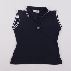 May include: Navy blue sleeveless polo shirt with white trim on the collar and armholes. The shirt features the "D&G" logo in white on the chest. The collar has a white stripe and the brand name "DOLCE&GABBANA" is visible.