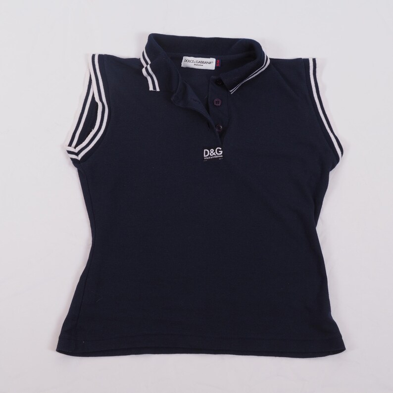 May include: Navy blue sleeveless polo shirt with white trim on the collar and armholes. The shirt features the "D&G" logo in white on the chest. The collar has a white stripe and the brand name "DOLCE&GABBANA" is visible.