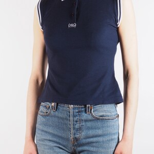 May include: Navy blue sleeveless polo shirt with white trim on the collar and armholes. The shirt features a three-button placket and the "D&G" logo. The shirt is paired with blue jeans.