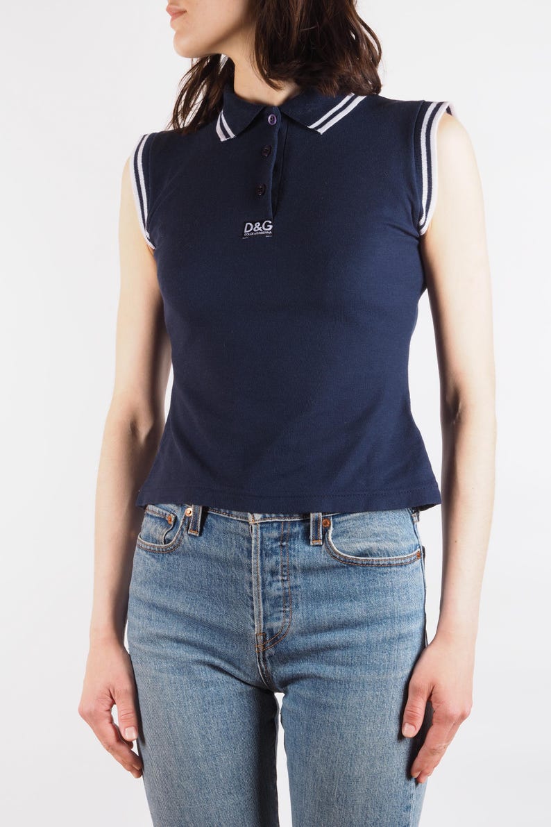 May include: Navy blue sleeveless polo shirt with white trim on the collar and armholes. The shirt features a three-button placket and the "D&G" logo. The shirt is paired with blue jeans.