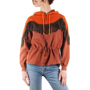 May include: A rust-colored hooded sweatshirt with a drawstring waist and a zip-up front. The top portion of the hoodie is orange, and the shoulders are adorned with brown fringe. The model is wearing blue jeans.