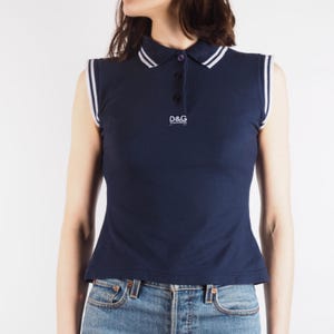 May include: Navy blue sleeveless polo shirt with white trim on the collar and armholes. The shirt features a three-button placket and the "D&G" logo. The shirt is paired with blue jeans.
