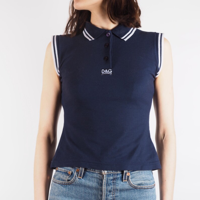 May include: Navy blue sleeveless polo shirt with white trim on the collar and armholes. The shirt features a three-button placket and the "D&G" logo. The shirt is paired with blue jeans.