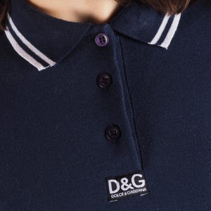 May include: Navy blue polo shirt with a white collar and white stripes. The shirt has three purple buttons and a D&G logo patch. The shirt is made of a soft, comfortable material.