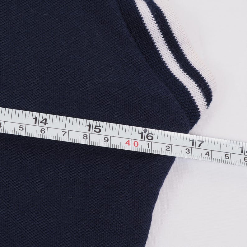 May include: Close-up of a navy blue polo shirt with white stripes on the collar. A measuring tape shows a measurement of approximately 16 inches across the chest. The shirt is made of a textured fabric.
