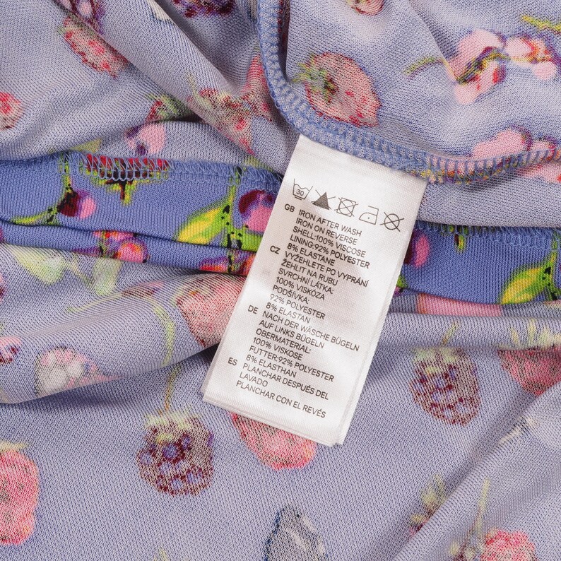 May include: Close-up of a light purple fabric with a floral pattern in pink and green. A white tag with care instructions is visible. The tag includes text in English, Czech, German, and Spanish.