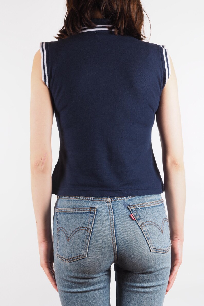 May include: Navy blue sleeveless top with white and light blue trim on the collar and shoulders. The top is paired with light blue denim jeans. The back view shows the details of the top and jeans.