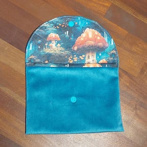 Puede incluir: Un bolso de mano turquesa con una escena de hongos caprichosa en la solapa. La solapa presenta una colorida ilustración de hongos y un paisaje de fantasía. El bolso de mano se cierra con un botón turquesa.