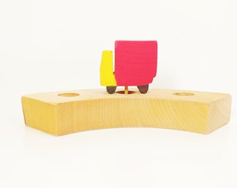 May include: A wooden toy truck with a red body and yellow cab sits on a wooden base with two holes. The truck is painted with simple details and has a small wooden dowel for a flagpole.