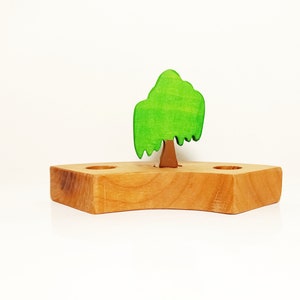 May include: A wooden tree with green leaves and a brown trunk, standing on a wooden base with two holes. The base is curved and has a natural wood finish.