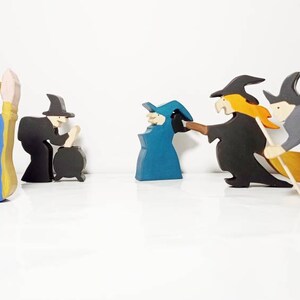 May include: A set of five wooden figurines depicting witches and a princess. The princess is wearing a blue and yellow dress and is holding a wand. The witches are wearing black and gray robes and hats. One witch is holding a broom, another is holding a cauldron, and the other two are flying.