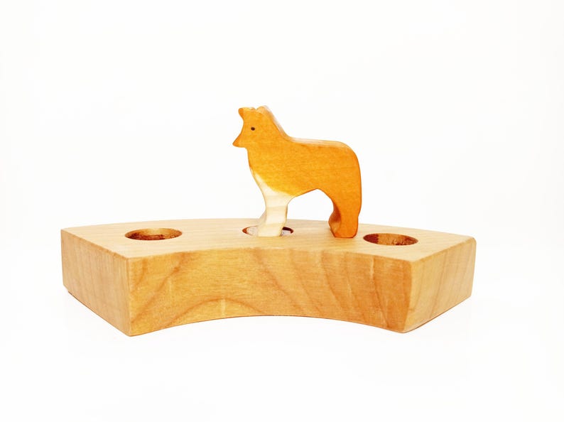 May include: Wooden toy set featuring a brown and white dog standing on a wooden base with two holes. The base is shaped like a crescent moon.