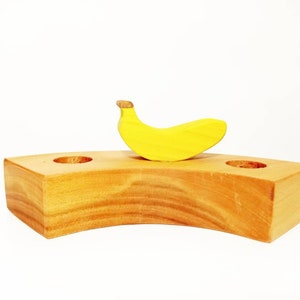 May include: A wooden banana holder with two holes for candles. The holder is shaped like a crescent moon and is made of light brown wood. A yellow wooden banana sits on top of the holder.