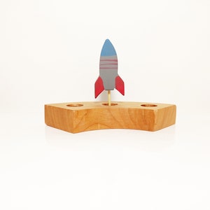 Könnte beinhalten: Ein hölzernes Raketenspielzeug mit roter und blauer Farbe, das auf einem Holzsockel mit drei Löchern steht. Der Sockel ist in Form eines Halbmondes.