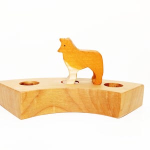 May include: Wooden toy set featuring a brown and white dog standing on a wooden base with two holes. The base is shaped like a crescent moon.