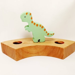 May include: A wooden dinosaur toy with orange spots stands on a wooden base with two holes. The dinosaur is green and the base is light brown.