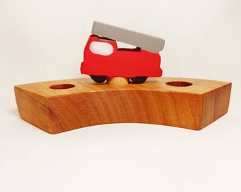 May include: A wooden toy fire truck with a ladder, painted red with black wheels, sits on a wooden base with three holes. The base is a light brown color.