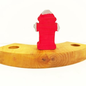 May include: A red wooden fire hydrant toy sits on a curved wooden base. The base has two holes on either side.
