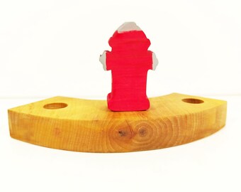 May include: A red wooden fire hydrant toy sits on a curved wooden base. The base has two holes on either side.