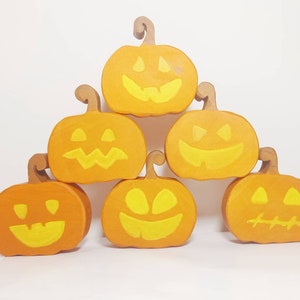 Può includere: Sei zucche di legno con facce dipinte di arancione e giallo. Le zucche sono disposte a forma di piramide.