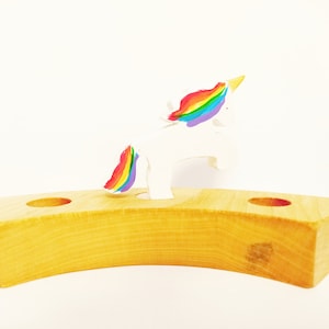 Puede incluir: Un portavelas de madera con un diseño curvo y dos aberturas circulares. Una figura de unicornio blanco con melena y cola de arcoíris se coloca encima. El soporte está hecho de madera de color claro.
