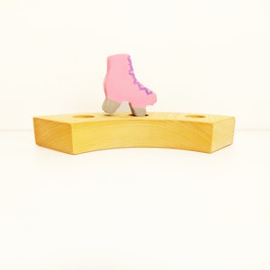 May include: A pink wooden roller skate toy sits on a light brown wooden base with two holes on either side.