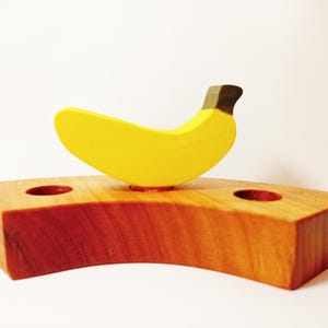 May include: A wooden banana shaped toy with a yellow body and a brown stem. The banana is sitting on a wooden base with two holes.