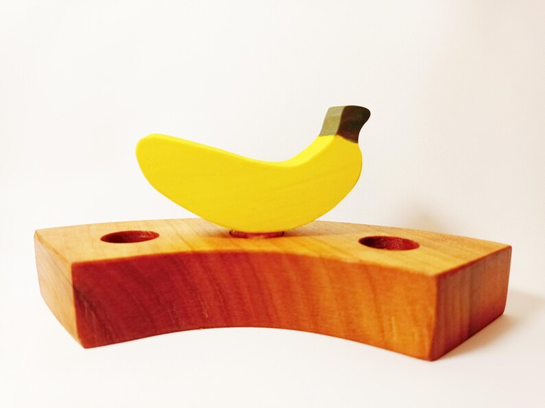 May include: A wooden banana shaped toy with a yellow body and a brown stem. The banana is sitting on a wooden base with two holes.