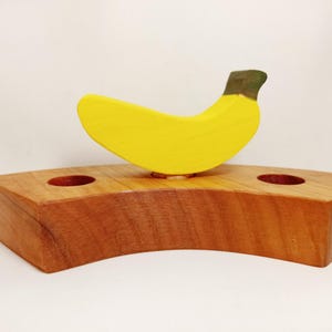 May include: A wooden banana shaped toy with a yellow body and a green stem sits on a wooden base with two holes. The base is brown and has a curved shape.